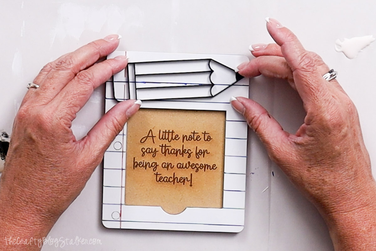 Hands gluing a pencil-shaped cutout onto a painted sticky note holder that reads "A little note to say thanks for being an awesome teacher!"