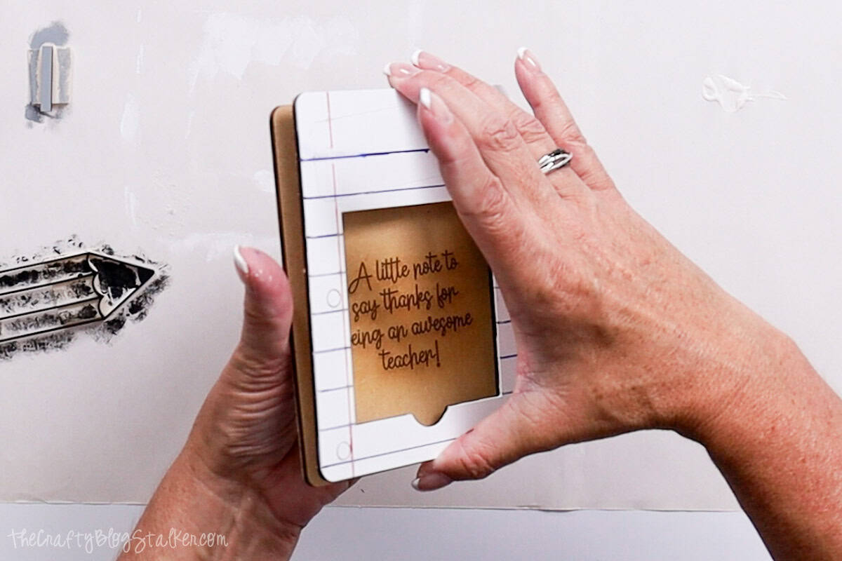 Hands assembling painted wood layers for a DIY sticky note holder shaped like notebook paper with the engraved message "A little note to say thanks."