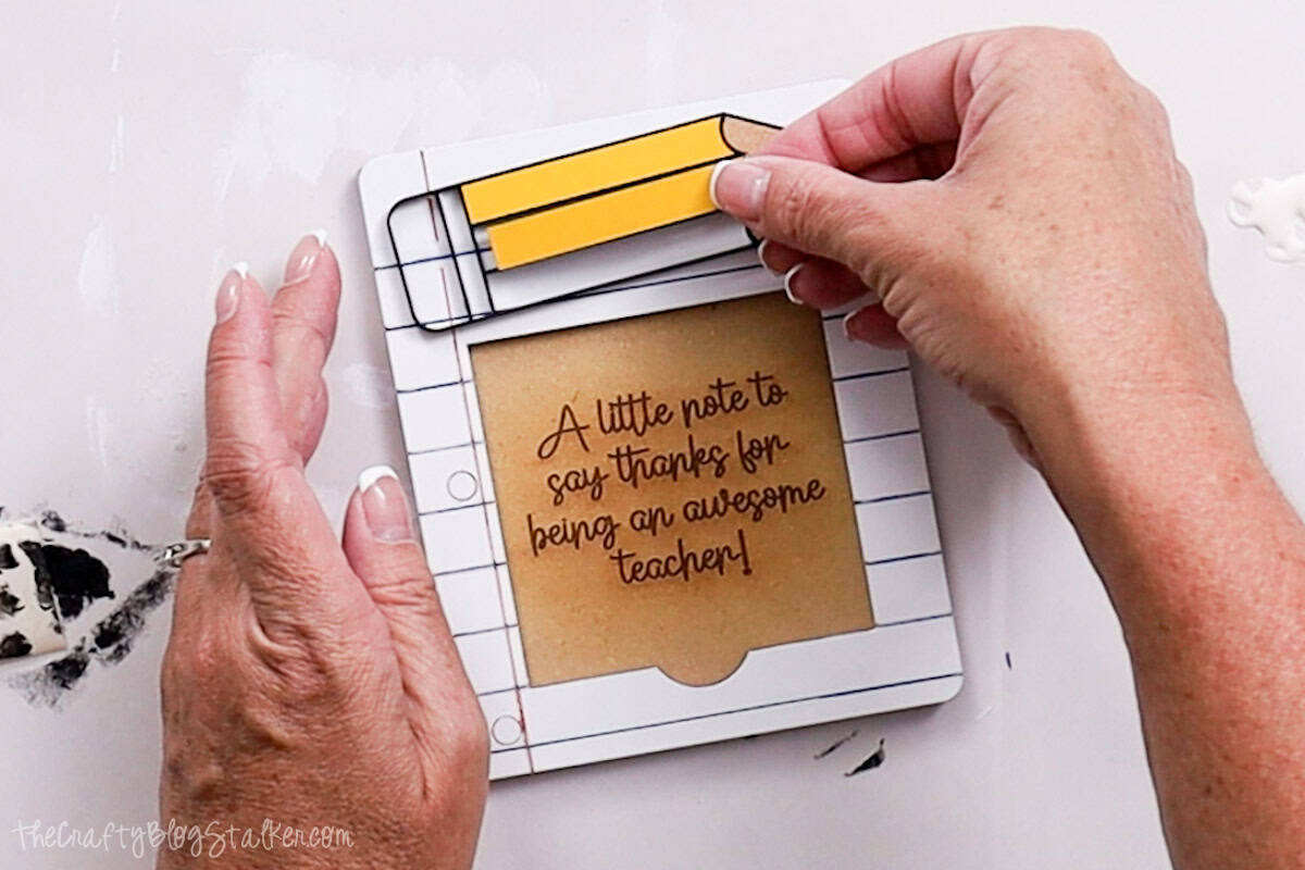 Hands gluing yellow pencil pieces onto a painted sticky note holder with the message "A little note to say thanks for being an awesome teacher!"
