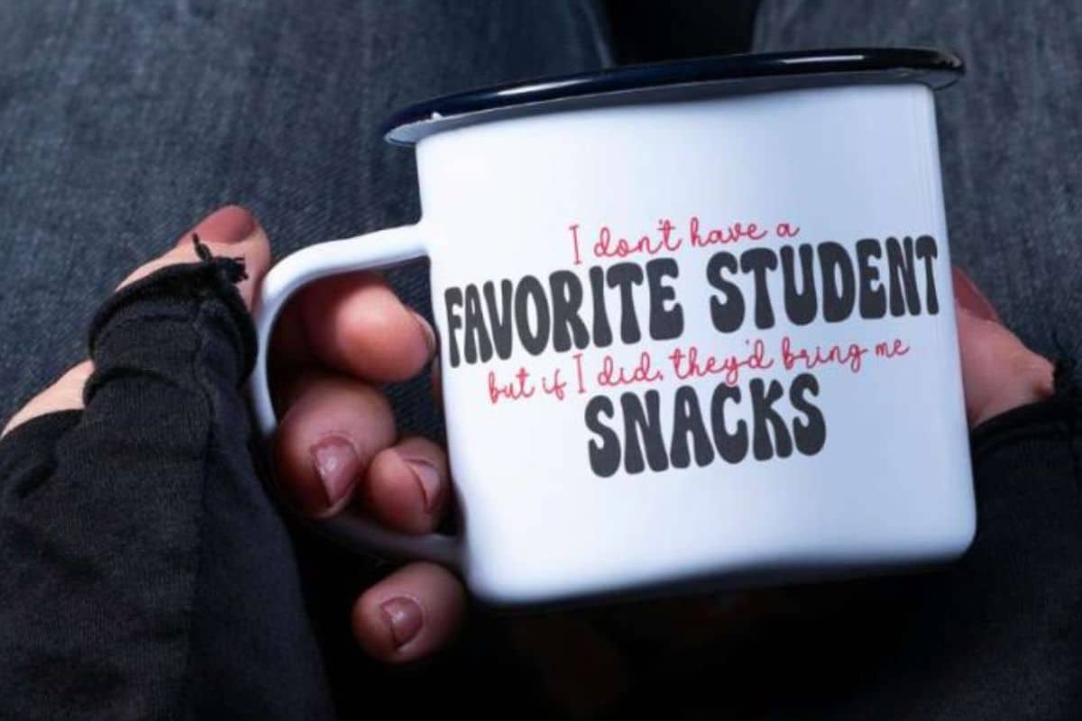 White enamel mug with black rim, held in hand, featuring a humorous teacher quote: "I don't have a favorite student but if I did, they'd bring me snacks."
