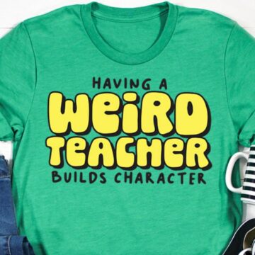 Green t-shirt with bold yellow and black text that reads "Having a weird teacher builds character," styled with jeans, sneakers, and a striped mug.