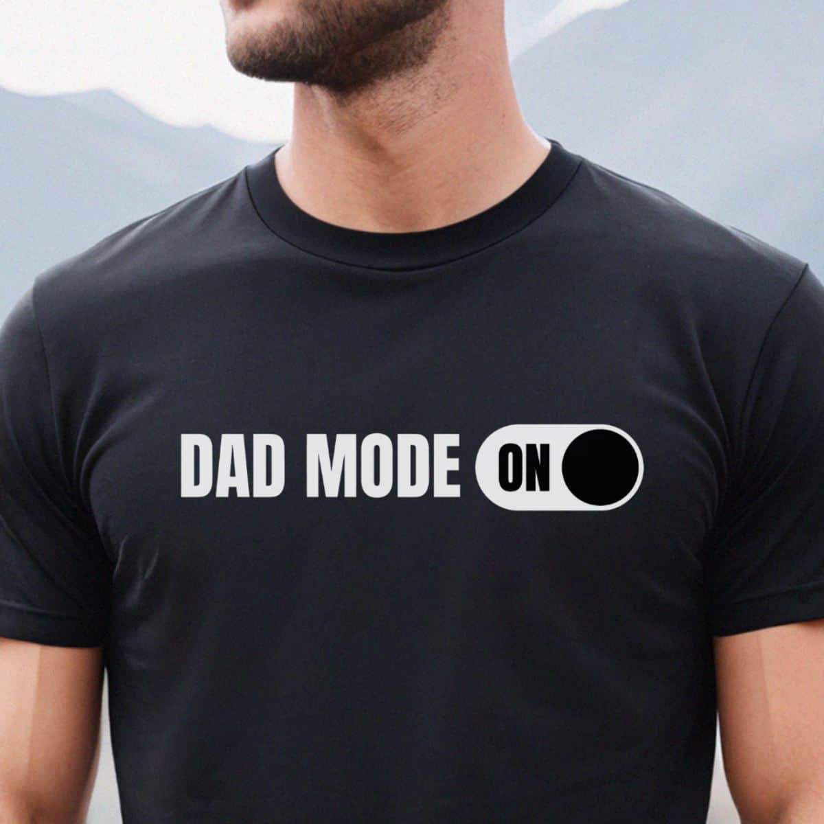 Black t-shirt with bold white text that reads "DAD MODE ON" next to a graphic switch flipped to the ON position, worn by a man outdoors.