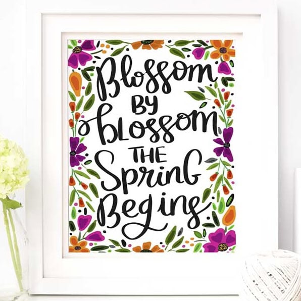 Framed spring quote print with colorful floral border reads "Blossom by blossom the spring begins" in whimsical black hand-lettered text on a white background.