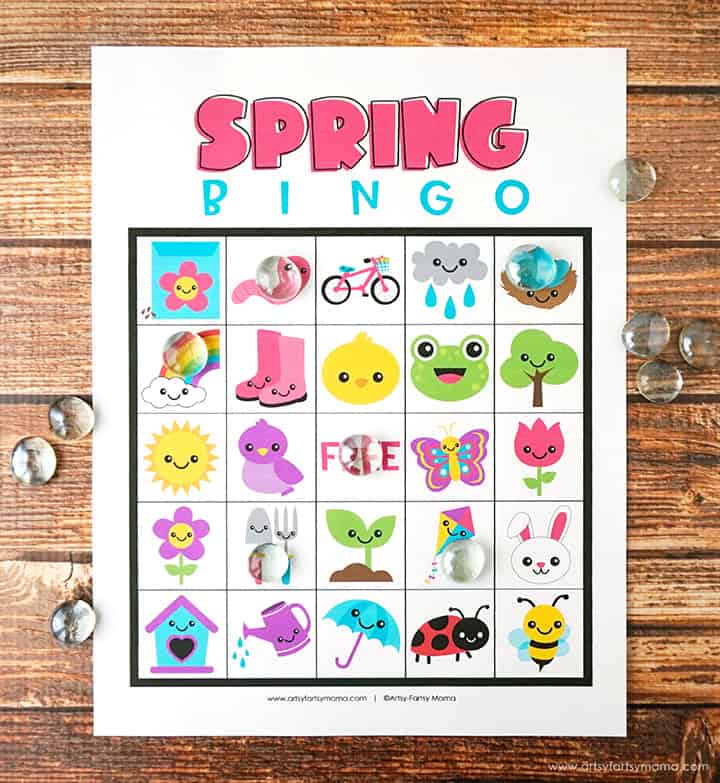 Printable spring bingo card with cute icons like flowers, frog, bunny, sun, umbrella, butterfly, and tree, placed on a table with glass markers.
