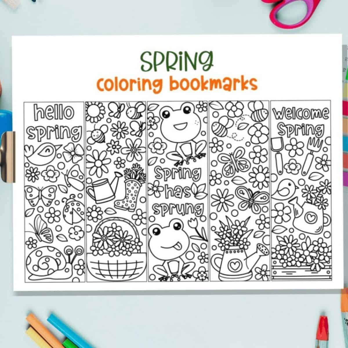 Printable sheet of spring-themed coloring bookmarks with drawings of flowers, frogs, birds, and gardening items, surrounded by craft supplies on a tabletop.