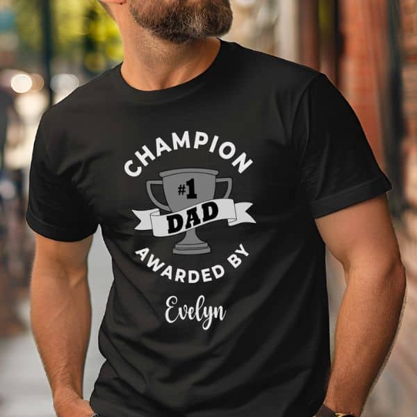 Man wearing a black t-shirt that says “Champion #1 Dad Awarded by Evelyn” with a trophy graphic, standing outdoors on a city sidewalk.