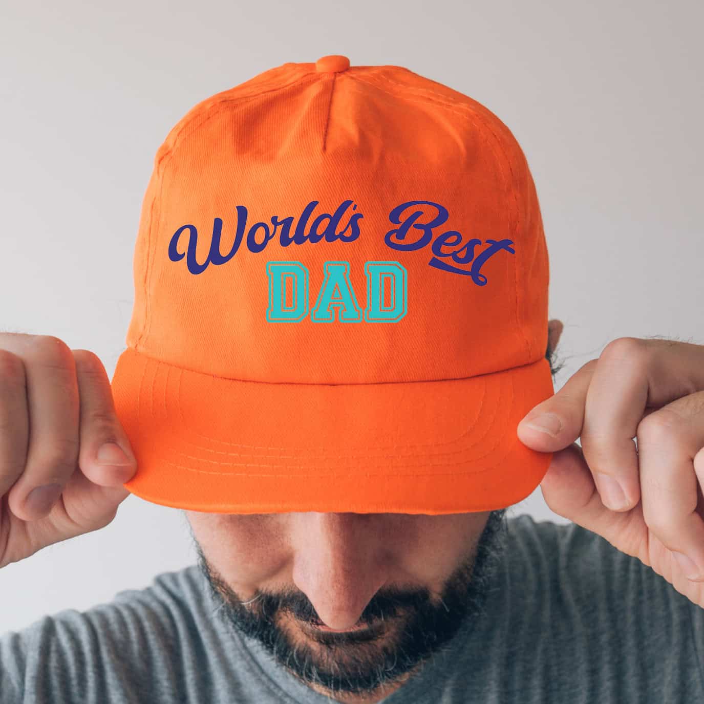 Man wearing a bright orange cap that says “World’s Best Dad” in blue and teal lettering, with his hands adjusting the brim.