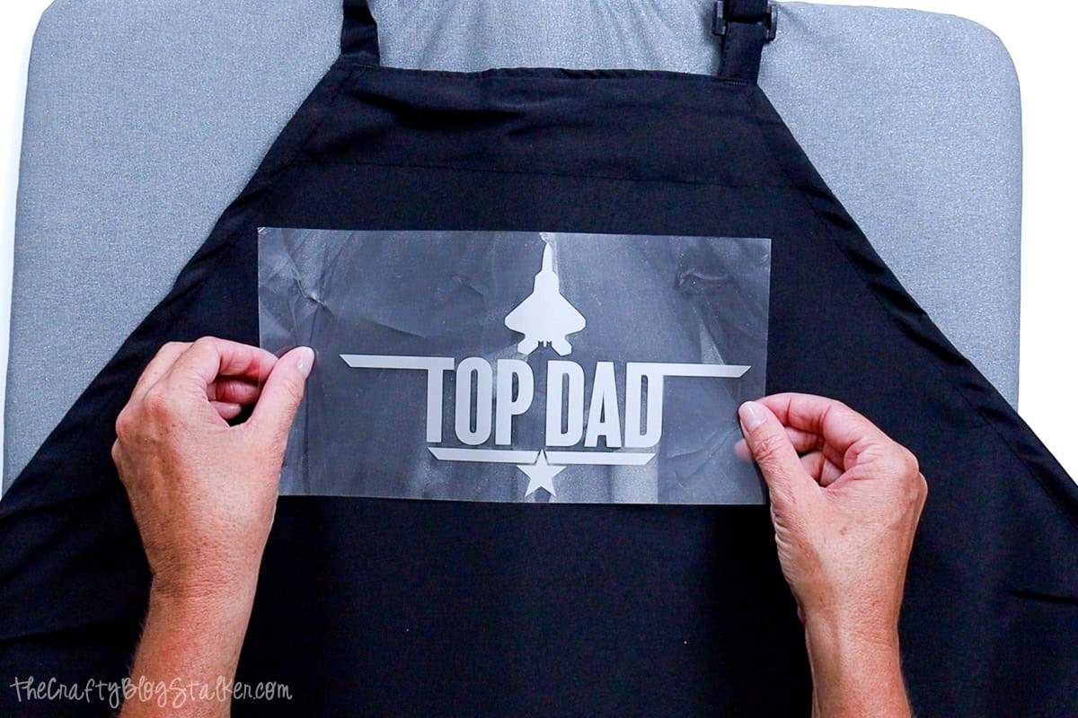 Hands positioning a silver “Top Dad” heat transfer vinyl design on a black apron, aligning it on a pressing mat for a DIY Father’s Day craft project.