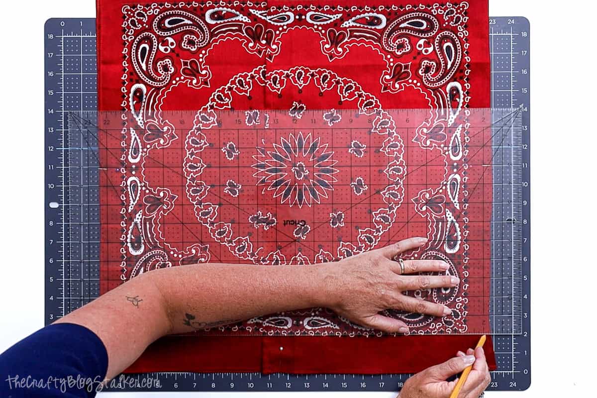 A person uses a clear quilting ruler and pencil to mark a 2-inch sewing line on red bandana fabric laid out on a cutting mat, prepping for a sewing project.