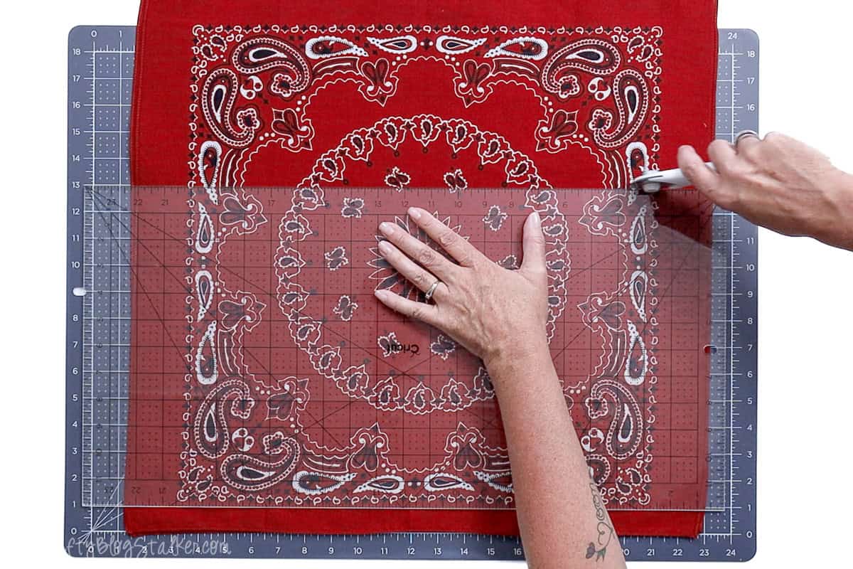 Hands cutting two red bandanas to 13 inches using a clear quilting ruler and rotary cutter on a gridded cutting mat, preparing fabric for a sewing project.
