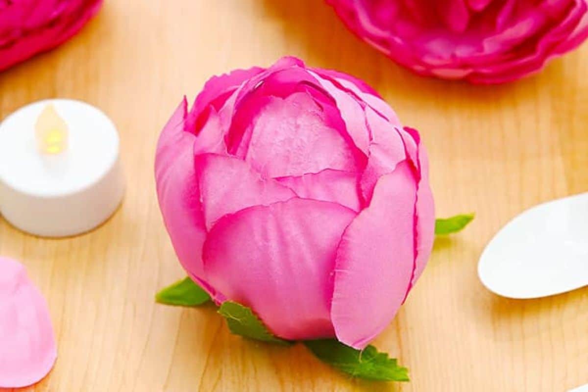 Bright pink artificial flower wrapped around a flameless tealight candle, displayed on a wooden surface with crafting tools and petals nearby.