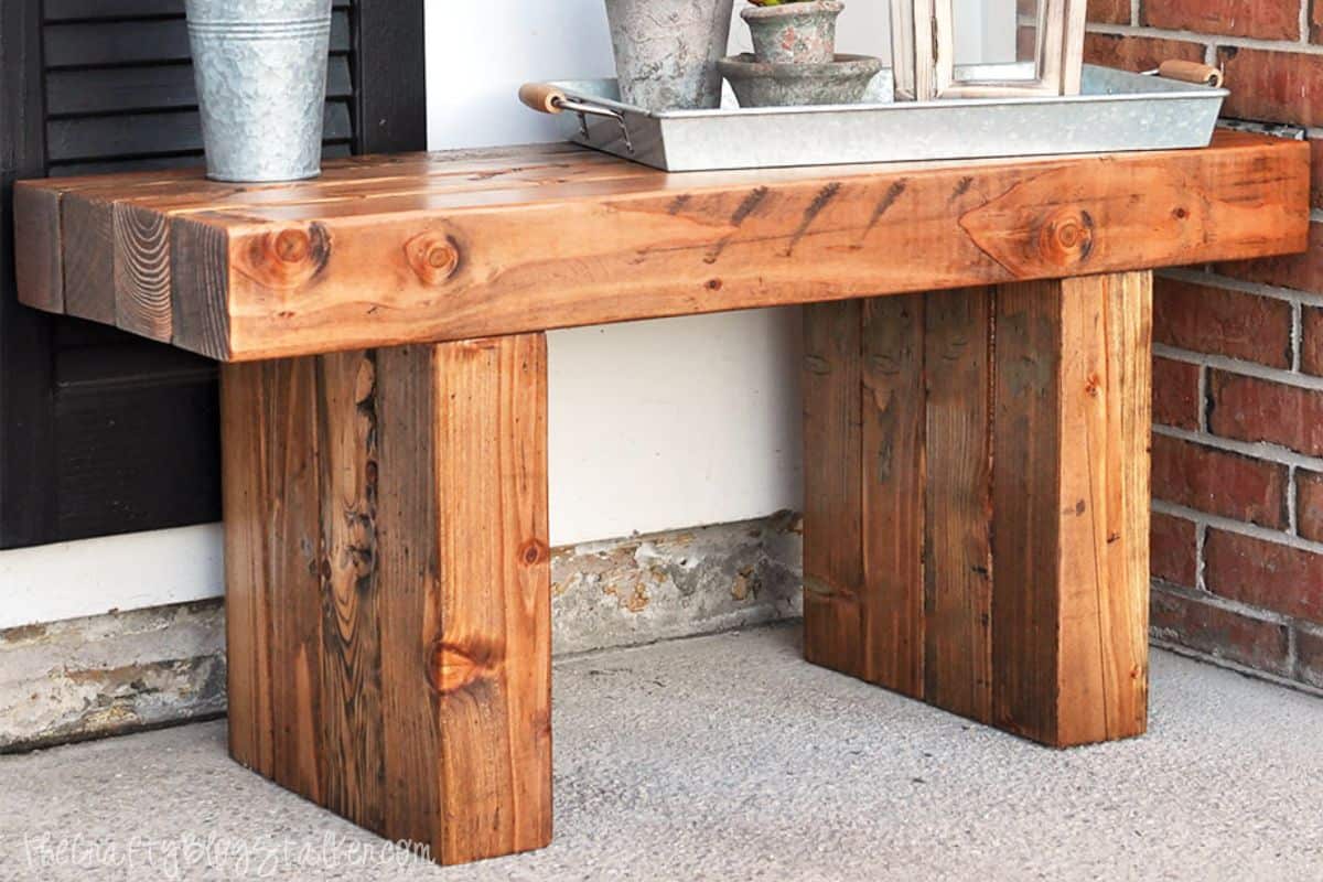 Finished wooden outdoor bench with a rich stained finish, styled on a porch with metal planters, a tray, and decorative accents.