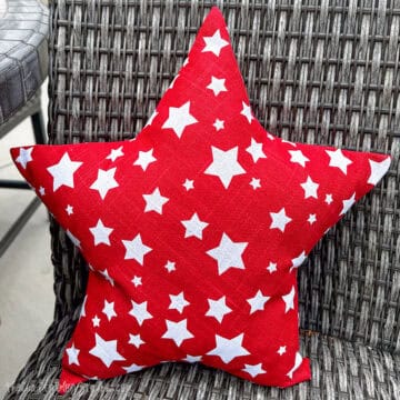 Red star-shaped throw pillow with white stars, resting on a woven wicker chair—perfect for patriotic or outdoor decor.