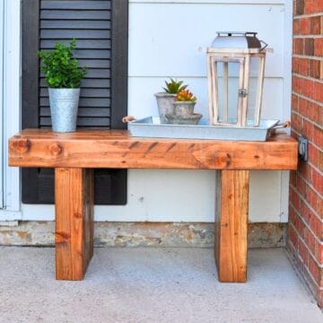 Rustic wooden bench on a porch, decorated with a potted plant, galvanized tray, succulents, and a white lantern against white siding and brick walls.