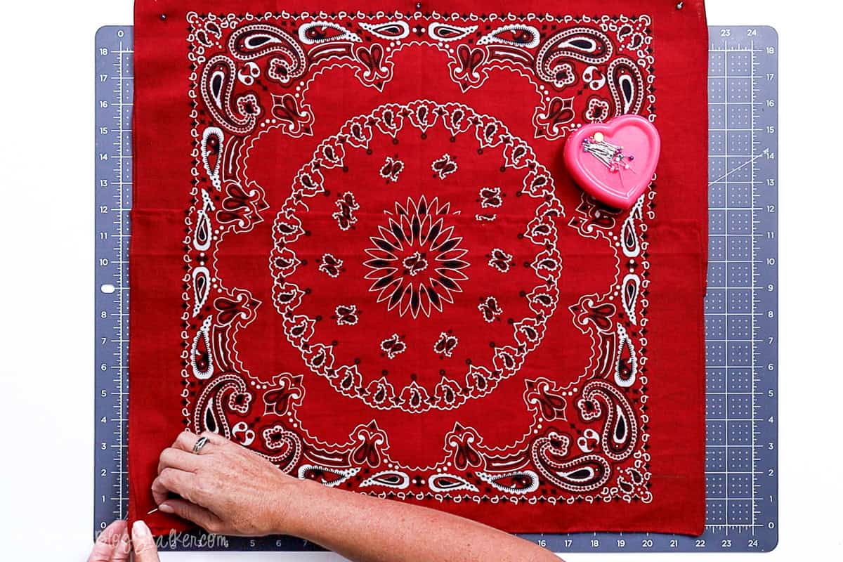 Hands pinning the corners of a red bandana square on a cutting mat, with a heart-shaped pin holder nearby, preparing fabric for sewing a DIY pillow cover.