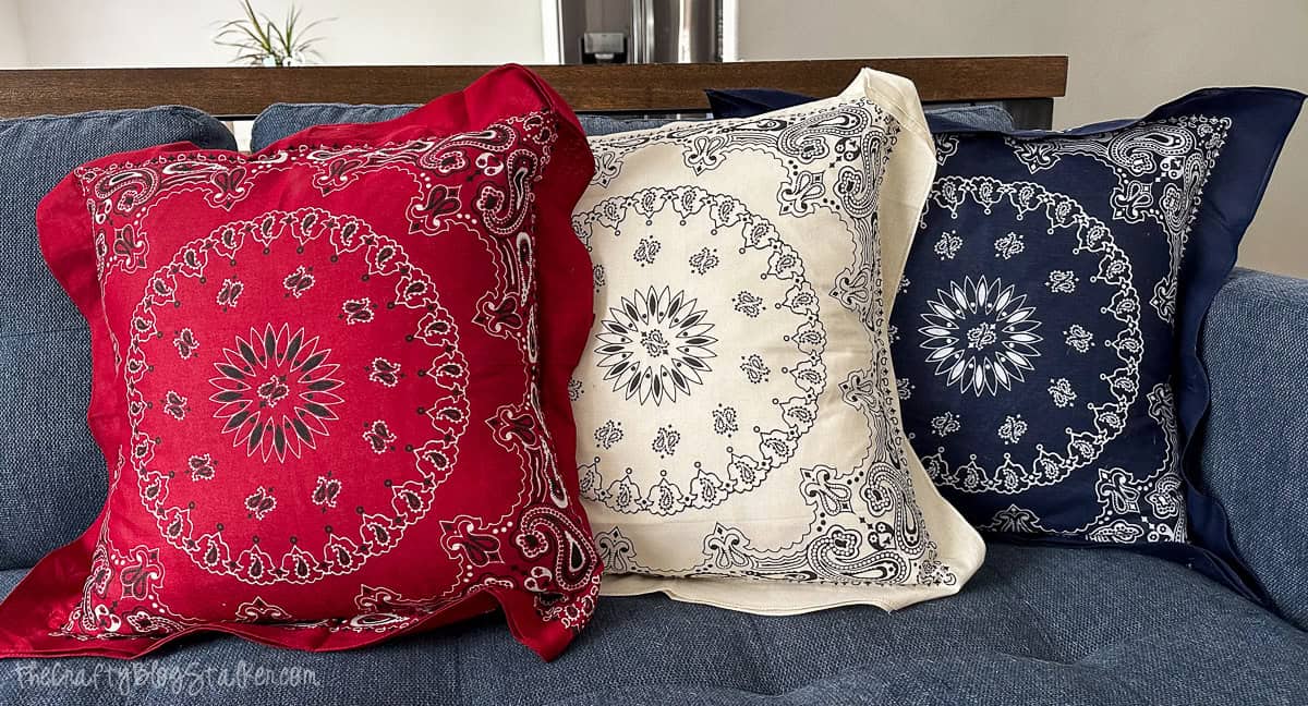 Three square bandana-style pillows in red, cream, and navy with paisley and mandala designs, displayed on a blue couch for a patriotic or western decor vibe.