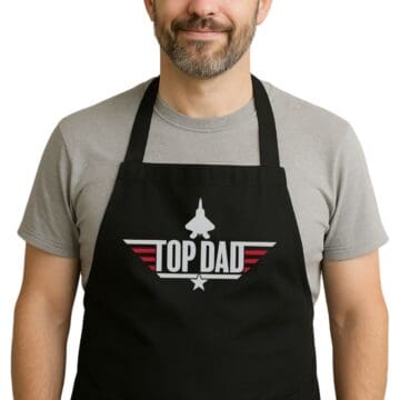 Man wearing a black apron with a fighter jet graphic and bold text that reads “Top Dad,” styled like the Top Gun logo, over a gray t-shirt.