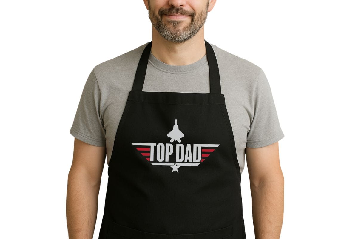 Man wearing a black apron with a "Top Dad" design featuring a jet, red stripes, and a star, inspired by the Top Gun logo, perfect for Father's Day grilling.