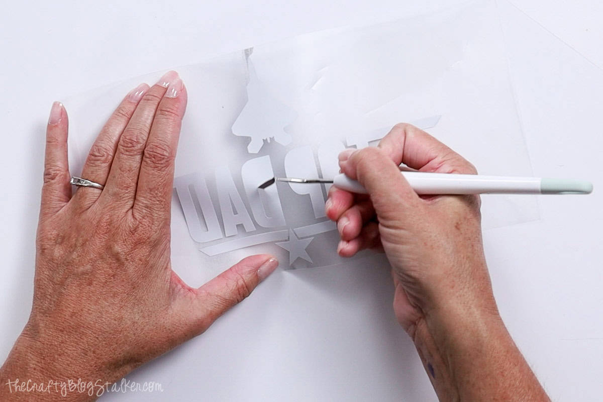 Hands using a weeding tool to remove excess vinyl from a mirrored "Top Dad" design, preparing the heat transfer graphic for a Father's Day apron craft.