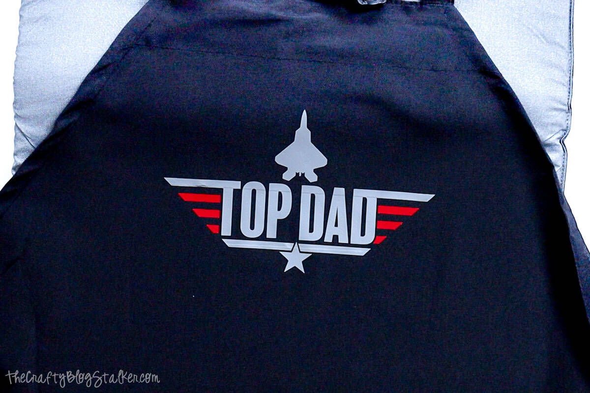 Black apron with a completed “Top Dad” heat transfer vinyl design in silver and red, featuring a jet and star, slightly wrinkled after pressing.