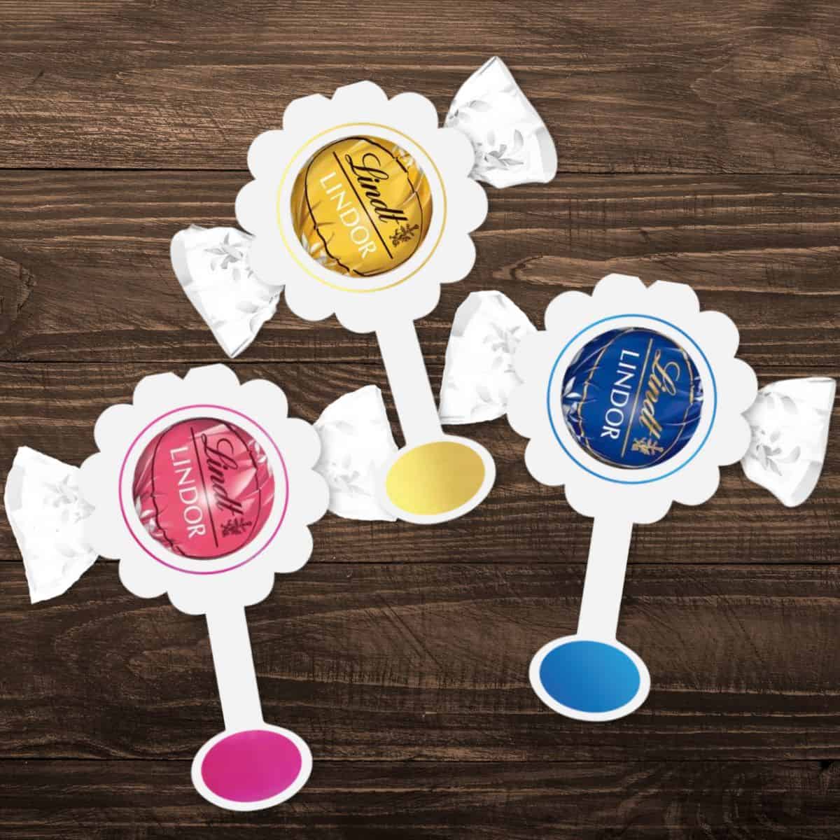 Three Lindor truffles wrapped in paper and framed in scalloped cardstock to resemble baby rattles, arranged on a wooden surface.