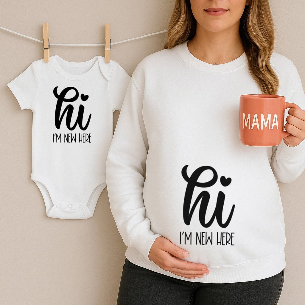 Pregnant woman wearing a sweatshirt that says "hi I'm new here," holding a "MAMA" mug, next to a matching baby onesie hanging on a line.