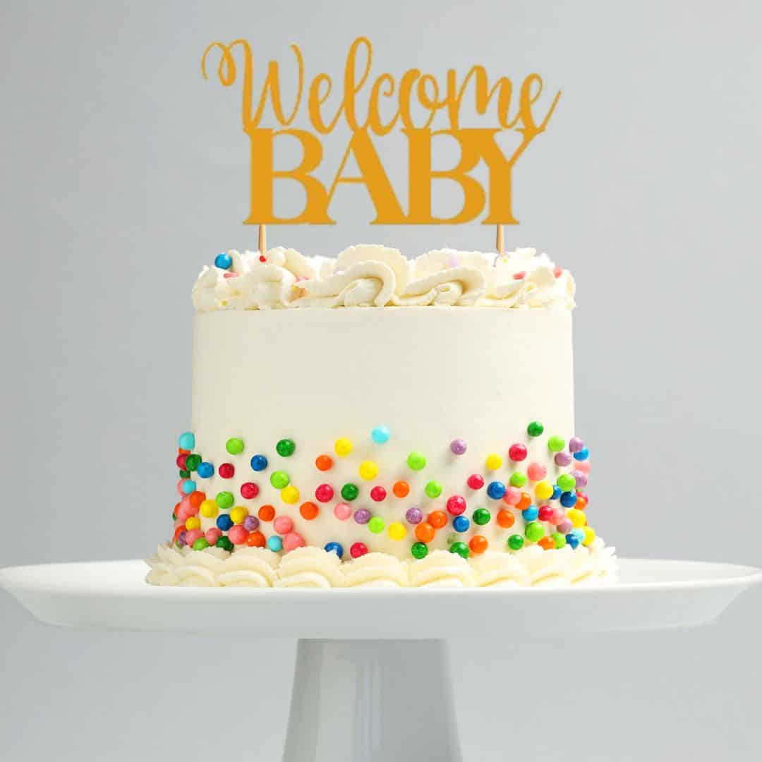 A white frosted cake topped with a yellow “Welcome Baby” cake topper and decorated with colorful candy sprinkles along the sides, perfect for a baby shower.