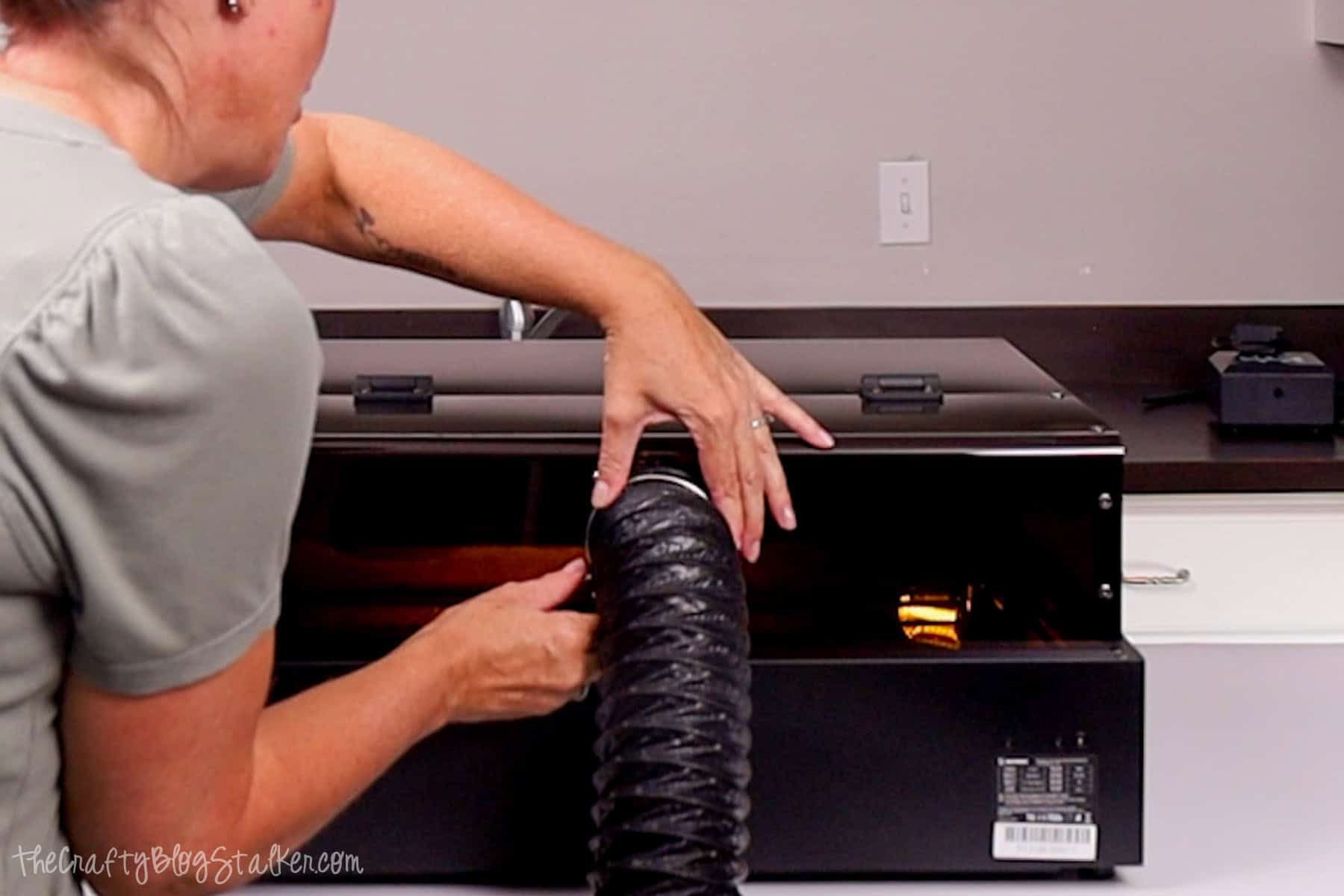 Person attaching a flexible black exhaust pipe to the back of the Acmer P3 Laser Engraver for ventilation setup in a home workspace.