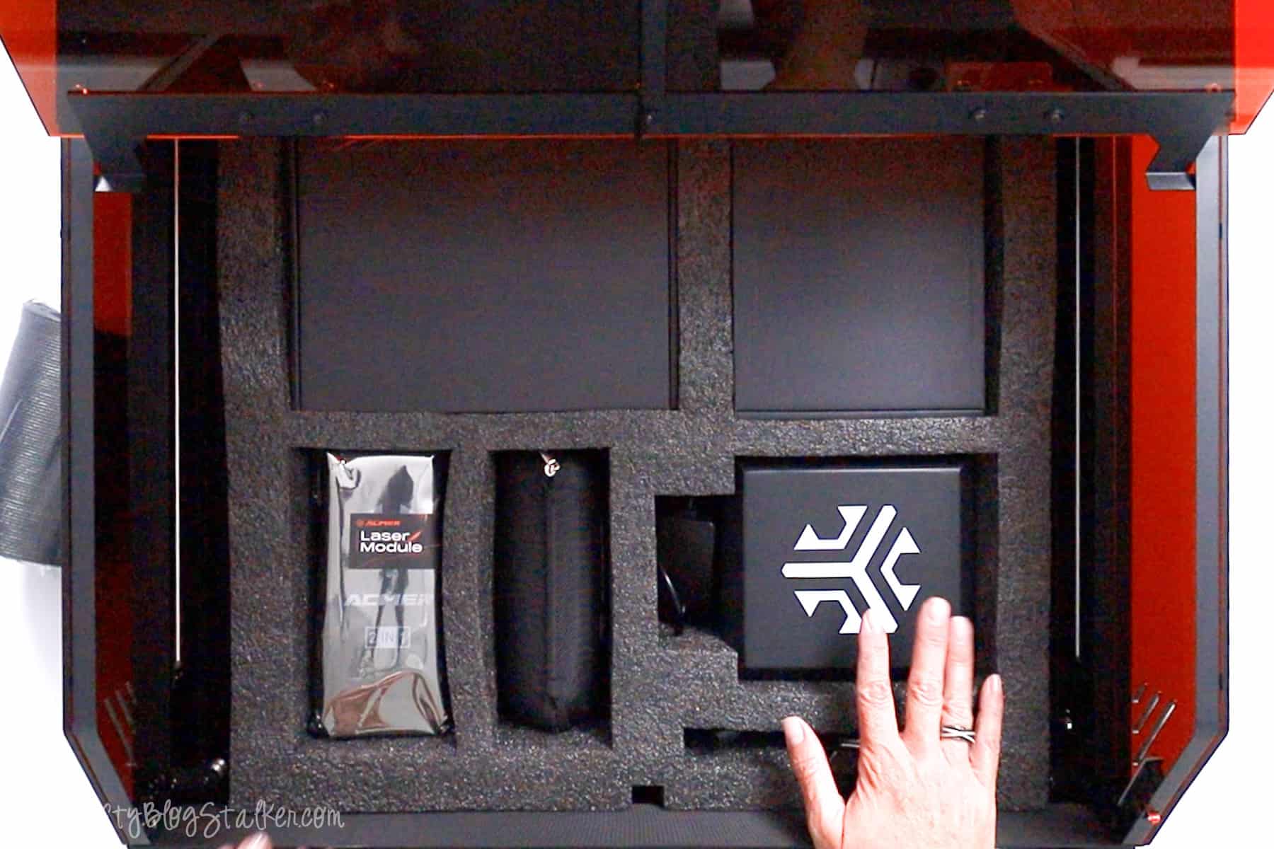 Interior view of the Acmer P3 Laser Engraver showing neatly packed components and accessories in foam, with a hand reaching toward the branded module box.