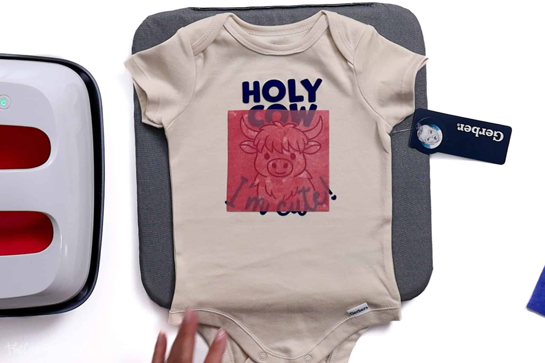 Red vinyl cow layer being centered and placed over the navy blue text on a beige baby onesie, ready for heat pressing with a Cricut EasyPress.