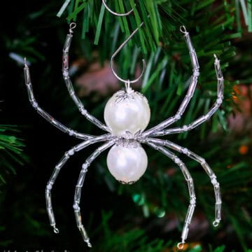 Beaded Christmas spider ornament with pearl body and silver wire legs, hanging from a green Christmas tree branch on a metal hook.