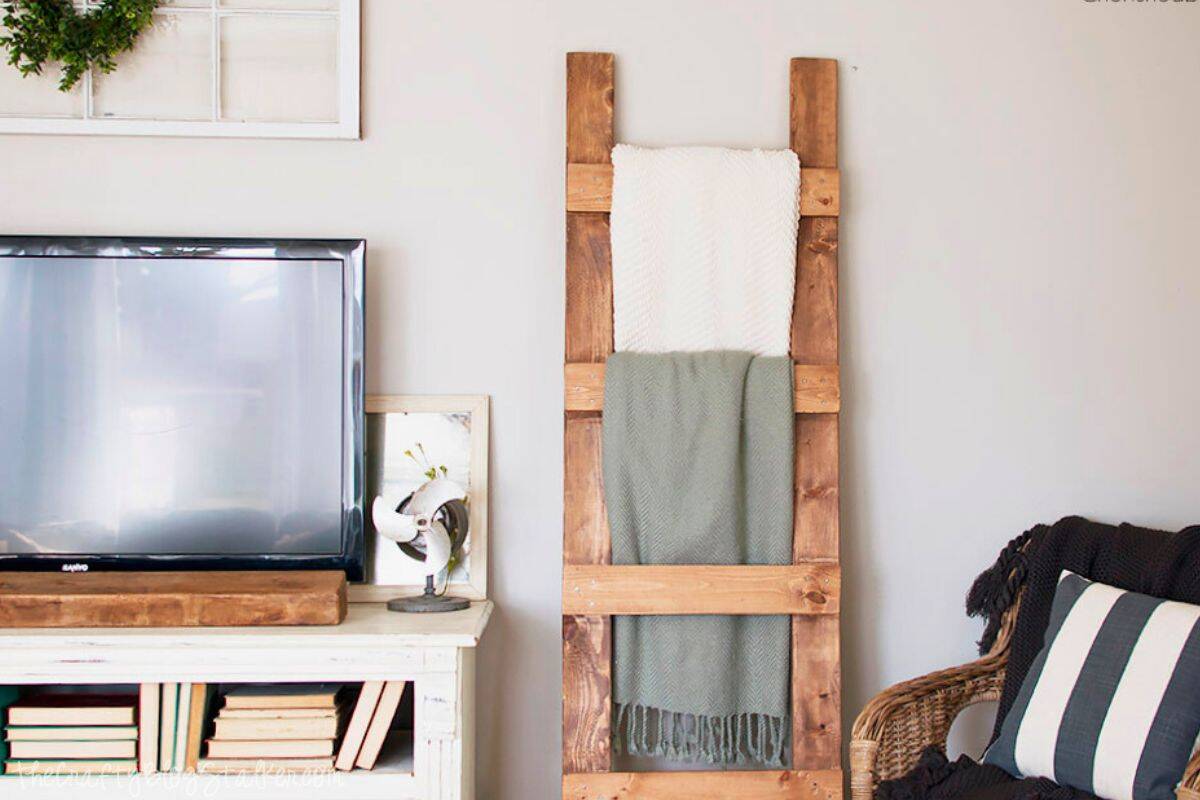Cozy living room corner with a wooden blanket ladder holding folded throws, next to a TV stand with books and a striped cushion on a wicker chair.