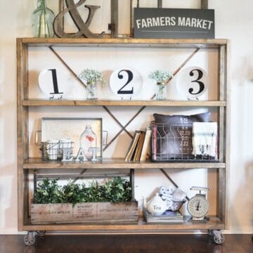 Rustic wooden bookshelf on casters styled with farmhouse decor, numbered plates, greenery, vintage books, crates, and a "Farmers Market" sign on top.
