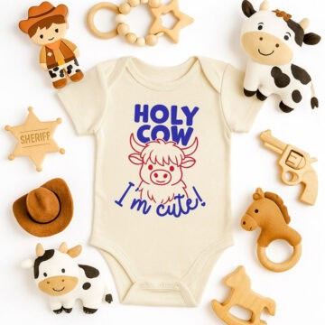 Cream baby onesie with “Holy Cow I’m Cute!” design, surrounded by plush cows, cowboy toys, wooden teether, sheriff badge, toy gun, and felt hat.