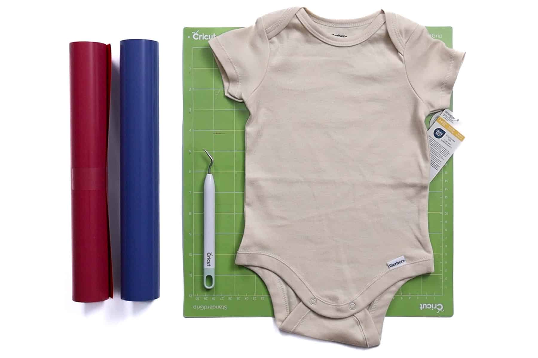 DIY baby onesie supplies including a blank onesie, red and blue heat transfer vinyl, Cricut weeding tool, and green Cricut cutting mat.