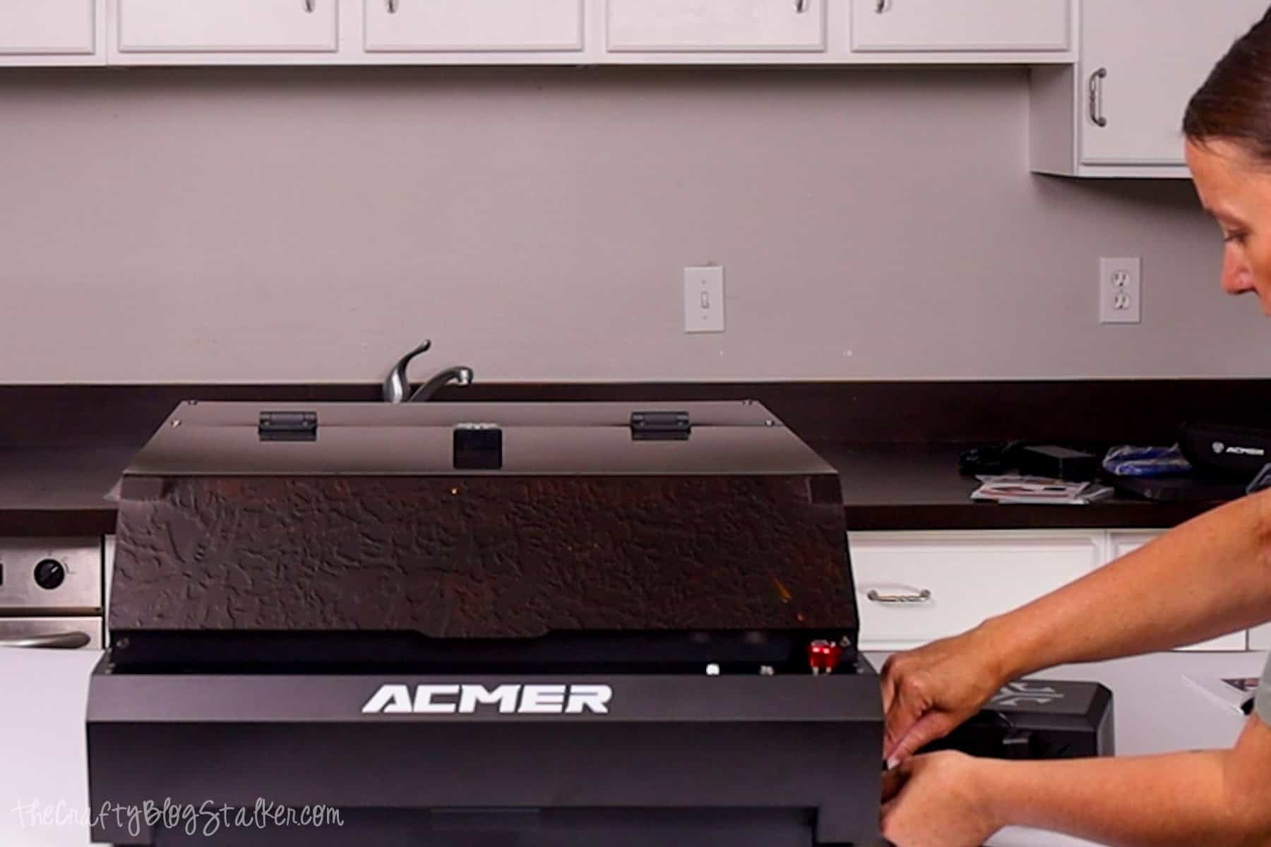 Person plugging in the Acmer P3 Laser Engraver on a countertop, preparing the machine for its first use with accessories nearby.