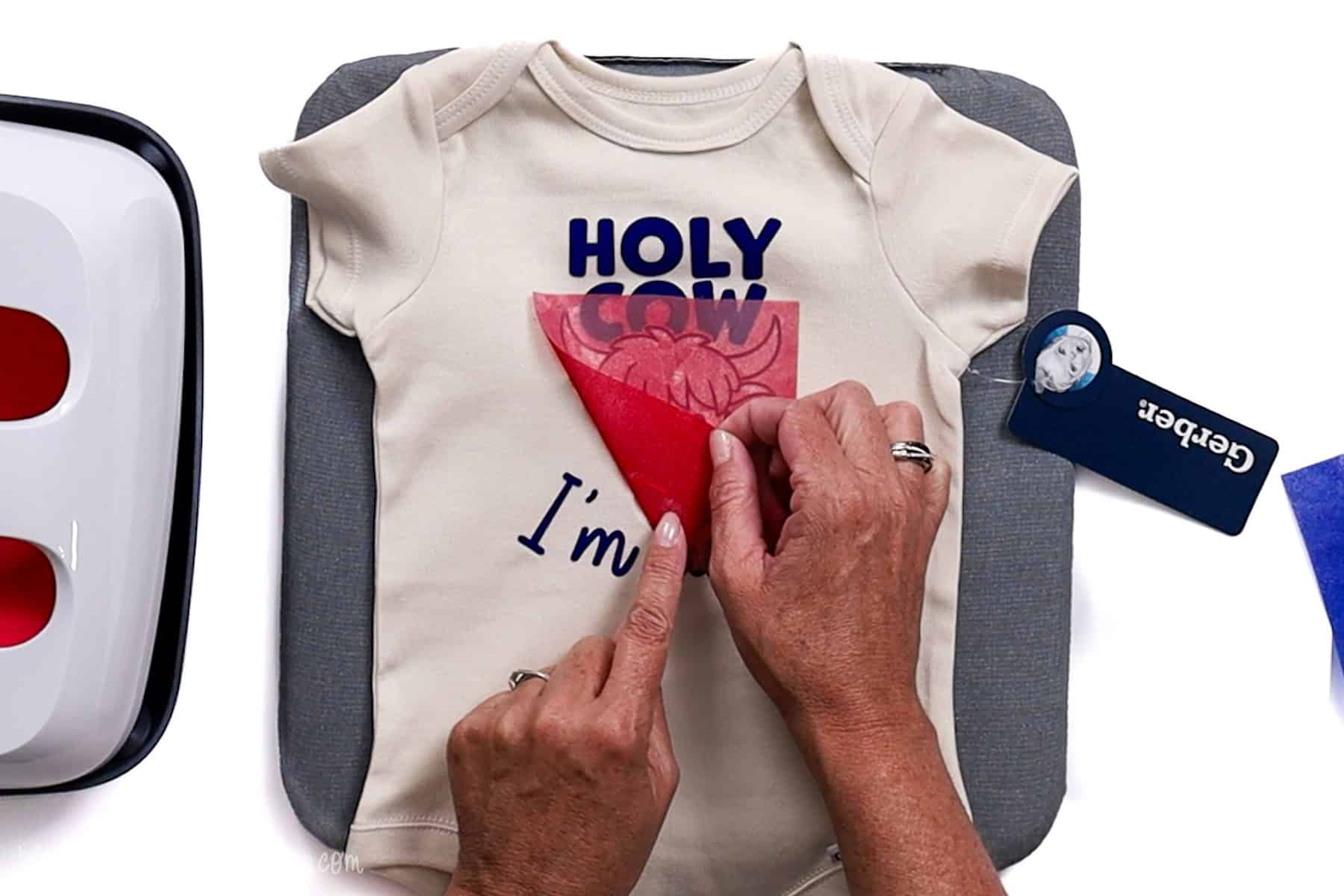 Person peeling red vinyl carrier sheet from a baby onesie and noticing part of the design hasn’t fully adhered after heat pressing with a Cricut EasyPress.