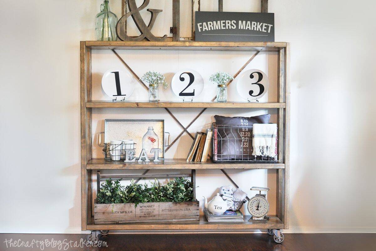 Rustic rolling bookshelf with wooden shelves and farmhouse decor, including vintage bottles, number plates, greenery, books, and a wire basket with blankets.