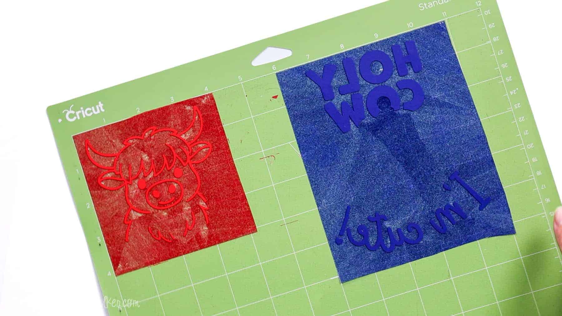 Red and navy blue flocked heat transfer vinyl designs weeded and ready on a Cricut cutting mat, featuring a cow graphic and text for a baby onesie project.