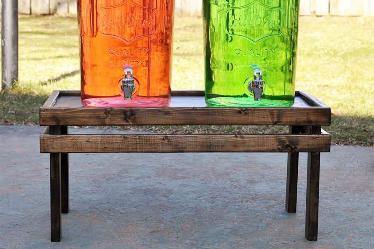 Two colorful drink dispensers, one orange and one green, sit on a rustic dark-stained wooden stand outdoors on a sunny day.