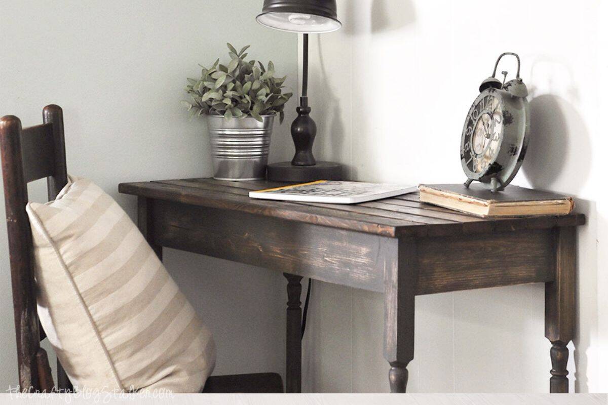 Rustic wooden writing desk in a cozy corner with a striped pillow on a chair, metal desk lamp, potted plant, vintage clock, and a couple of books on top.