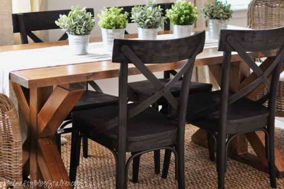 Rustic dining room with a wooden X-brace farmhouse table, black cross-back chairs, and small potted plants as a centerpiece on a neutral woven rug.