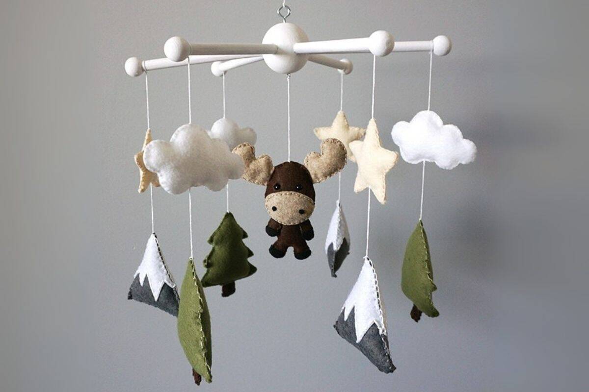 Felt woodland baby mobile with a moose, trees, stars, clouds, and snow-capped mountains hanging from a white wooden frame.