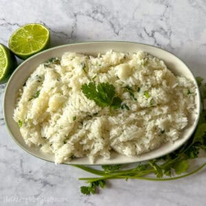 A bowl of fluffy cilantro lime rice garnished with a fresh cilantro sprig, placed next to halved limes and cilantro stems on a marble surface.