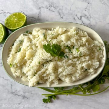 A bowl of fluffy cilantro lime rice garnished with a fresh cilantro sprig, placed next to halved limes and cilantro stems on a marble surface.