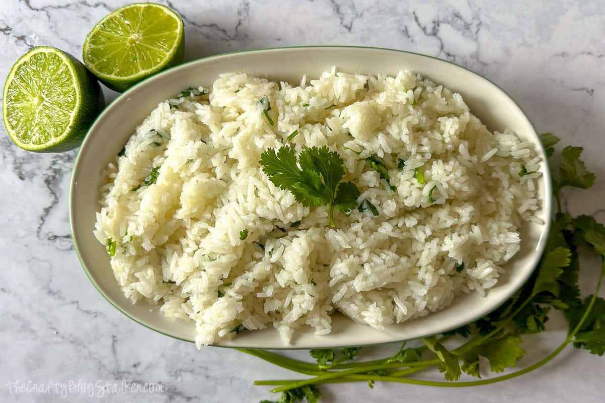 A bowl of fluffy cilantro lime rice garnished with fresh cilantro, served with halved limes on a marble countertop.