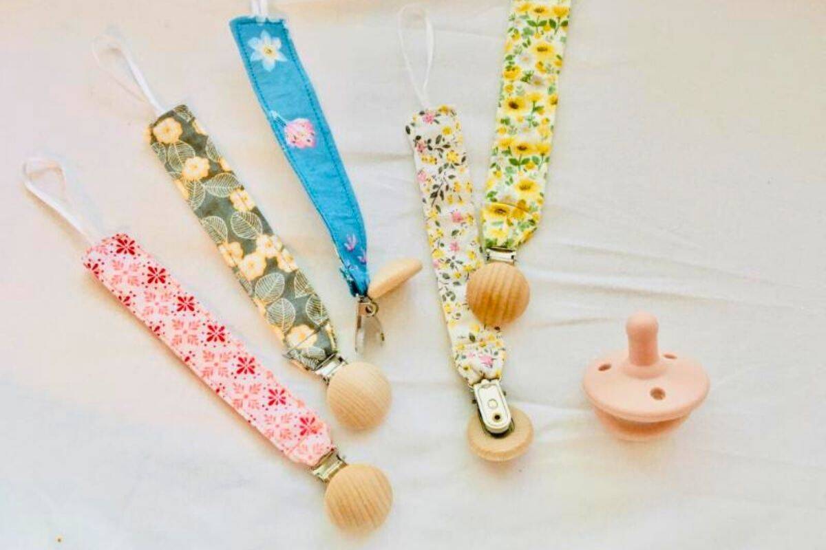 Five handmade fabric pacifier clips with colorful floral prints and wooden clips, shown beside a pink pacifier on a white background.