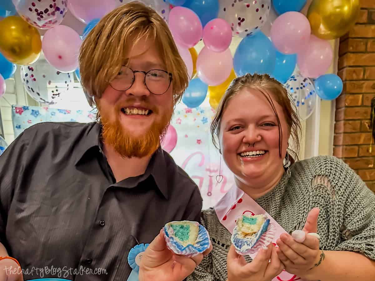 A smiling couple at a gender reveal party holding cupcakes with blue filling, surrounded by pink, blue, and gold balloons.