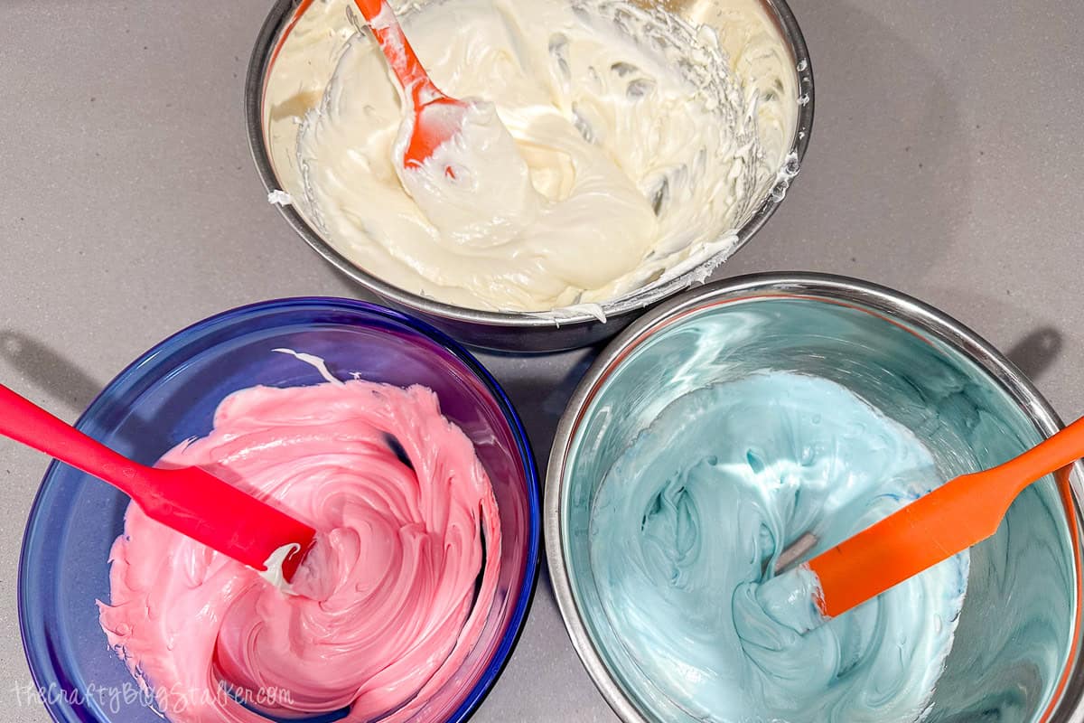 Three bowls of frosting in white, pink, and blue with spatulas, prepared for decorating gender reveal cupcakes.