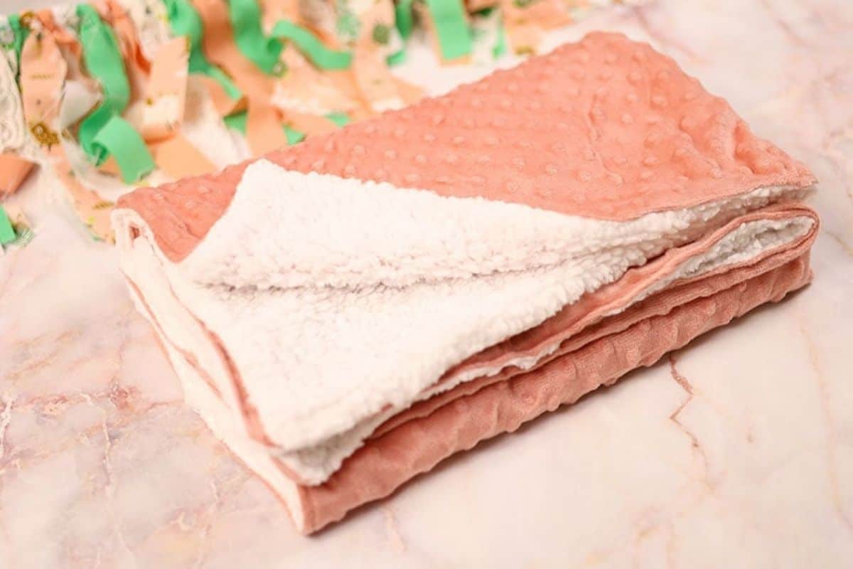 Folded baby blanket with a soft pink minky fabric on one side and white sherpa fleece on the other, displayed on a marble surface.